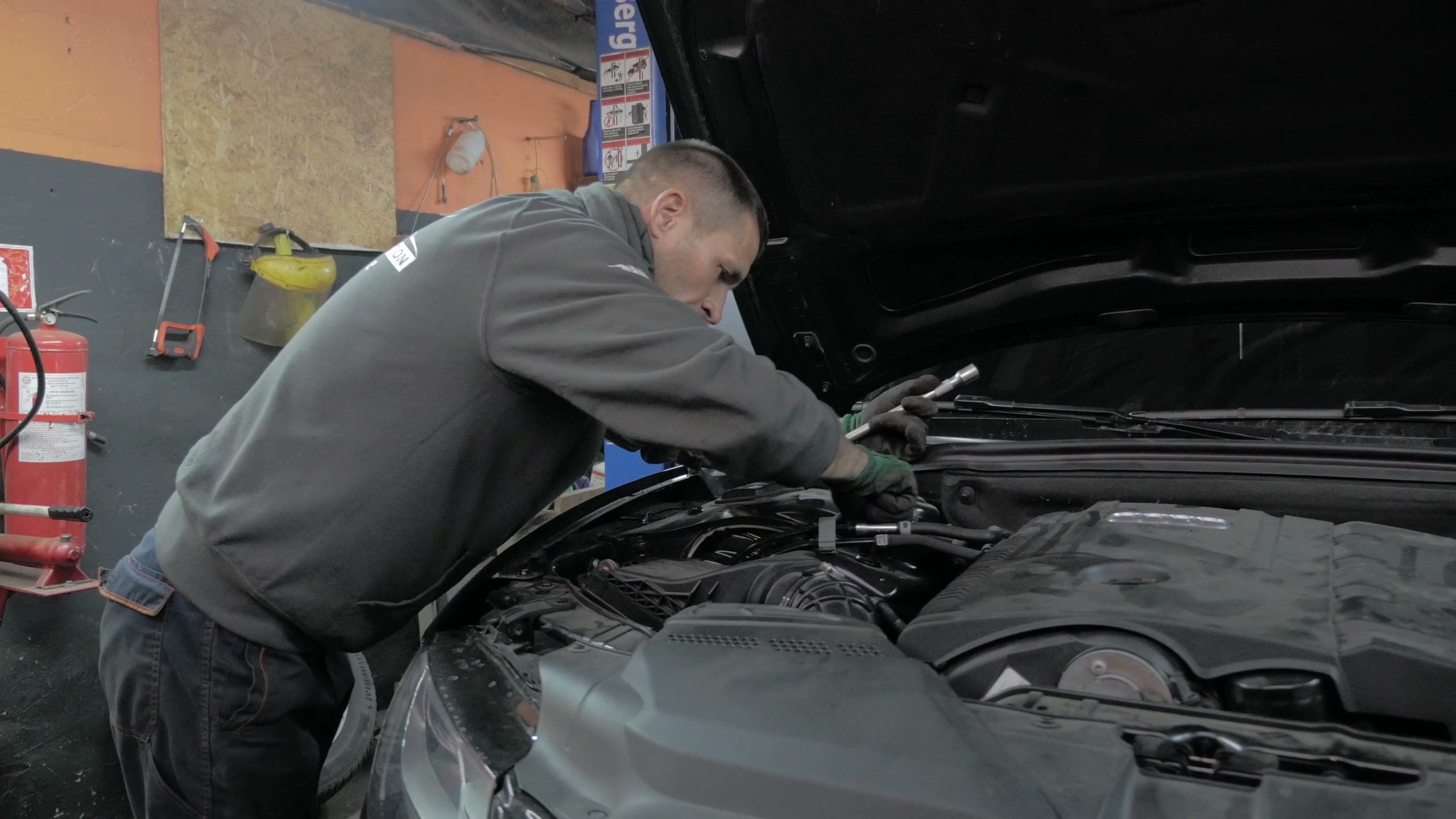 A mechanic in a grey sweatshirt leaning into the open engine bay of a dark car, a socket wrench in his gloved hand